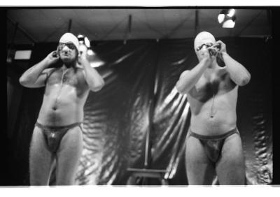 Brian Routh and Martin von Hasselberg stand next to each other while securing swim caps to their heads. They wear goggles and jockstraps. A tarp hangs behind them.