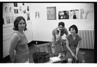 Three people are situated in a gallery setting. On the right, a person sits on a loveseat bench and looks at the person standing to their right. They stand with their hands behind their back and a clip holds their bob-shapped hair away from their face. Behind them is a person who rests their right hand to their chin and regards the camera.
