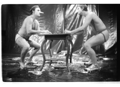 Brian Routh and Martin von Hasselberg crouch at two ends of a table. They wear shower caps, nose prosthetics, jock straps, and dress shoes. 