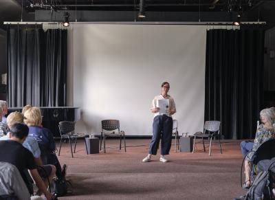 A speaker stands in front of a blank projector screen in the Grand Luxe Hall, addressing a crowd. She clutches papers and a phone with both hands. She wears navy pants, a white top, white sneakers, and glasses. Her hair is dark and slicked back. 
