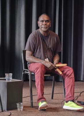 An aboriginal man with short-cropped hair sits on a chair, holding two thick, wooden sticks in his hands. He wears red pants, a brown t-shirt, neon green shoes, and a lanyard around his neck.
