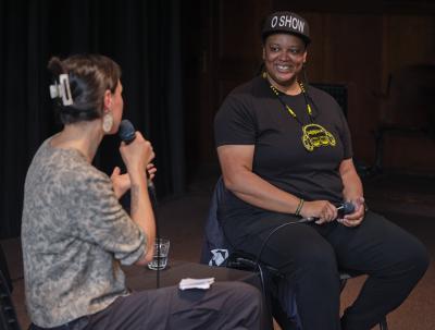 An interviewer and an artist sit side by side. The interviewer appears to be asking the artist a question, gesturing with their hands as they speak. The artist’s grin is wide and toothy. 
