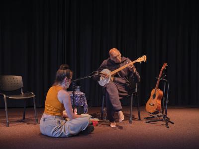 Shahzad Ismaily plays banjo while seated. Julia Úlehla sits cross-legged on the ground, facing him. An empty chair, a guitar, and a blue-clothed table can be seen in the background. 