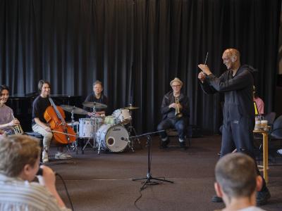 A bald conductor stands in the Grand Luxe Hall, surrounded by a group of musicians seated in a circle around him. The conductor wears all black, smiling as he waves his baton. The group features percussion, brass, and string instrumentalists. 
