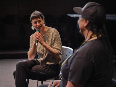 A person wearing a paisley button down, dark pants, and black earrings holds a microphone with both hands, smiling as they speak to an artist sitting across from them.
