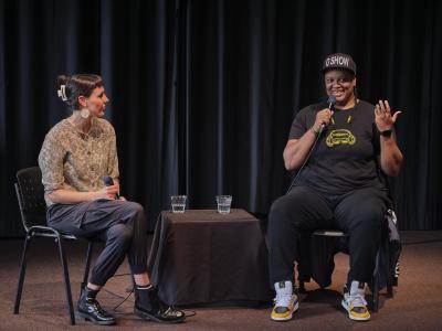 An interviewer and an artist sit side by side, addressing a crowd. The artist wears all black with pops of yellow, smiling as they speak into a microphone. The interviewer wears a paisley button down and crosses their legs, listening attentively.
