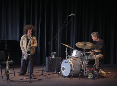 A saxophonist wears her hair in an afro and is dressed in a tan cardigan and black pants. Leaning forward with her eyebrows furrowed and eyes shut in concentration, she embodies the music while jamming with a grey-haired drummer.
