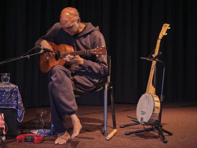 Shahzad Ismaily sits cross-legged, looking down while playing ukulele. A banjo on a stand and a wooden flute are arranged on the floor nearby. To his right, a red tambourine rests on the edge of a table covered in an ornate blue cloth.