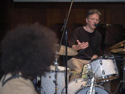 A white drummer with gray hair and tan pants sits behind a drum kit, gesturing with his hands as he speaks into a microphone. In the foreground of the image, a person with an afro is seen from behind. 
