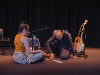 Wearing a dark tracksuit, Shahzad Ismaily kneels down on the floor, using one hand for balance and the other to arrange small square cards in an arching row. Julia Úlehla wears an orange top and sits facing them, cross-legged. Chairs, microphones, a small table, a guitar, and a banjo are arranged behind them.