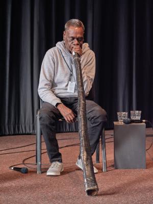An aboriginal man with short-cropped hair sits on a chair as he plays the didgeridoo. He wears black jeans, a gray hoodie, gray sneakers, and a lanyard. The didgeridoo appears to be covered in black duck tape. Several microphones lay untouched around him.
