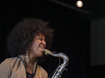 Side-profile view of a Black saxophonist with an afro wearing a tan top and cowrie shell earrings against a black background. The musician’s concentration is visible on her face as she bites down on her reed, scrunching her nose with furrowed eyebrows and closed eyes.
