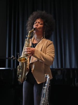 A Black saxophonist with an afro tilts her head back slightly as she plays, her tan cardigan in stark contrast against the black curtains behind them. Her eyebrows are furrowed and her eyes are shut as she vigorously blows air into her instrument. 
