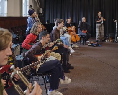 A group of around ten musicians sit in a semicircle in the Grand Luxe Hall. The group features string, woodwind, and brass musicians. The camera is focused on the side profiles of three young guitarists.
