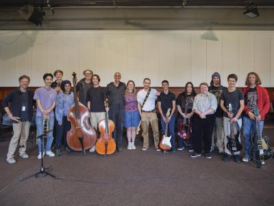 A group of diverse musicians stand in the Grand Luxe Hall, posing for a group photo. Some stand with their instruments while others pose with their hands clutched together or wrapped around their bandmates’ backs.
