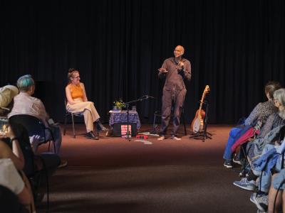 Shahzad Ismaily stands while speaking into a microphone, addressing the audience. Julia Úlehla sits to his left next to a small blue-clothed table. To his right is a ukulele and a banjo are propped up on black stands. 