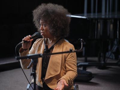 A Black person with a small afro and a tan cardigan speaks into a microphone, thoughtfully addressing a crowd. 

