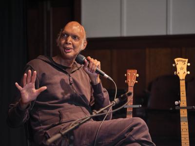 Shahzad Ismaily gestures with his hand as he speaks into a microphone, addressing a person off-screen. He wears a dark tracksuit. The fretboards of two stringed instruments can be seen in the background.