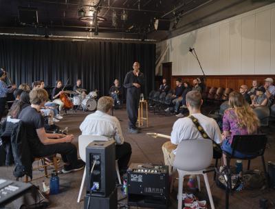 A group of musicians sit in a circle in the Grand Luxe Hall, facing a bald instructor wearing all black. The instructor stands in the middle of the circle, scratching his chin. The group features string, woodwind, drum, and percussion musicians. 
