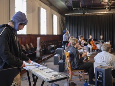 A person wearing a blue knit hat and a navy jacket stands as they play the xylophone. In front of them, a group of musicians are seated in a semicircle, mostly out of focus. The group features brass, percussion, and string instrumentalists. 
