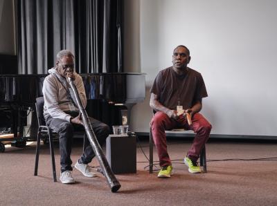 Two aboriginal Yolngu artists sit next to each other in the Grand Luxe Hall. The man on the right plays the didgeridoo while the man on the left creates rhythms with two short, thick wooden sticks. The didgeridoo player wears a gray hoodie, black jeans, and gray sneakers. His bandmate wears a brown t-shirt, red pants, and neon green sneakers.
