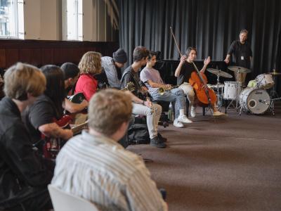A group of musicians sit in a semicircle, with a drummer at the farthest end. Next to the drummer sits a cellist who holds their hands up as they address the group. The rest of the instrumentalists pay careful attention to the cellist’s words. 
