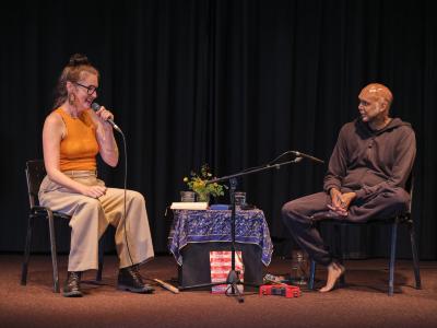 Julia Úlehla and Shahzad Ismaily are seated across from each other on a stage. She speaks into a microphone addressing Shahzad and wears an orange top, large hoop earrings and black glasses. Between them is a bouquet of yellow flowers and several knick knacks on a table covered in ornate blue cloth.
