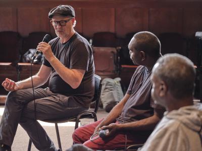 A man in a newsboy cap sits cross-legged as he speaks into a microphone. He wears a black t-shirt, gray pants, and glasses. White stubble decorates his features. Two men watch him as he speaks, facing away from the camera.
