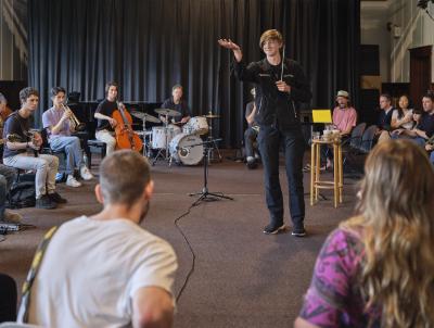 A young person dressed in all black stands in the center of the Grand Luxe Hall, holding a conducting baton in one hand and holding the other up to the sky. Around them sit a circle of various musicians, including a drummer and multiple string instrumentalists. 
