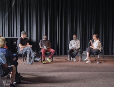 A panel of four speakers faces an audience, each holding a microphone. To the far right, a woman sits cross-legged, addressing the other three. An aboriginal man wearing black jeans and a gray hoodie is seated to her left, followed by another aboriginal man wearing red pants and a dark t-shirt. To the far left a white man wearing a newsboy cap sits cross-legged.