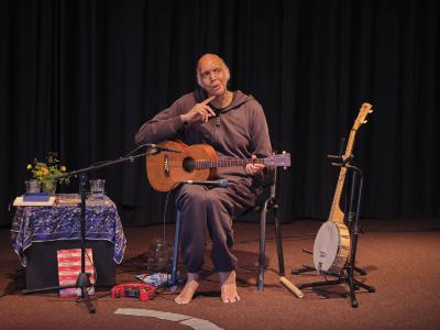 Wearing a dark tracksuit, Shahzad Ismaily sits on a chair with a microphone and a ukulele in his lap. A banjo is propped up on a stand to his left and a bouquet of yellow flowers on a small table covered in blue ornate cloth is to his right. The artist’s right index finger is pointed at his chin as he gazes to the left mid-thought.
