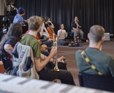 A group of musicians sit in a semicircle in the Grand Luxe Hall. The group features string, brass, and woodwind instrumentalists. Roughly half of the musicians sit with their backs to the camera. To the far end of the semicircle, a person wearing a striped sweater sits on the ground, addressing the rest of the group.
