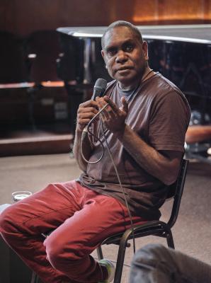 An aboriginal man with short-cropped hair sits in a black chair as he speaks into a microphone. He laces the microphone's wires through his fingers as he speaks. He wears red pants, a brown t-shirt, and a lanyard around his neck. 

