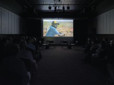 An audience sits in the darkness of the Grand Luxe Hall, facing a screen. On the screen is the image of an aboriginal man wearing a blue t-shirt and a yellow and black cap while leaning against a fence. He gazes at the natural landscape before him. The image is captioned I’m a Wägilak man.  
