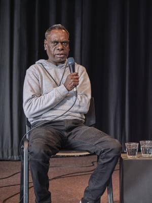 An aboriginal man with short-cropped hair holds a microphone up to his mouth with one hand and tucks the other beneath his arm. He wears black jeans, a gray hoodie and a lanyard around his neck. 
