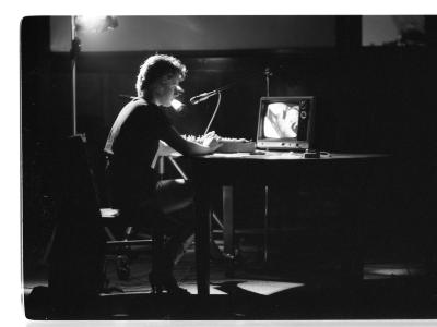 A television screen is set on top of a desk. The right-side profile of a person sat on a chair. They hold their hands together on the desks' surface and with an attentive expression, address the screen in front of them. A microphone standing behind the desk points downward at the person’s mouth. 
