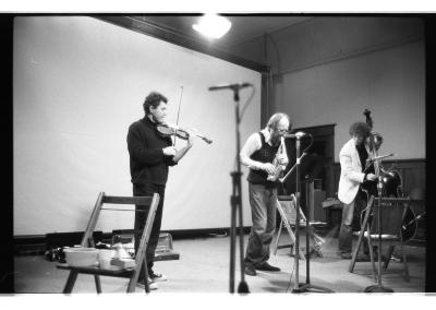 A black-and-white photo of David Prentice playing violin, Bill Smith playing soprano saxophone, and David Lee playing cello. They stand in a line in the Grand Luxe Hall. Microphones and chairs are set up around the performance space. A projector screen is pulled down behind the musicians. 