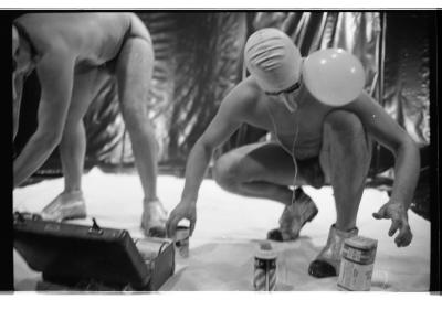 Brian Routh and Martin von Hasselberg crouch on a white tarp in the Grand Luxe Hall. Von Haseslberg wears a swim cap, goggles, and a prosthetic nose. A balloon is attached to his ear. Cans of shaving cream and a suitcase of other materials are positioned on the floor as well. 