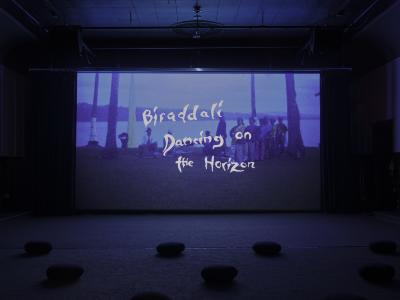 A blue screen with superimposed text reading Biraddali Dancing on the Horizon. A vast empty room is lined with chairs and ovular flat cushions placed carefully on the floor.