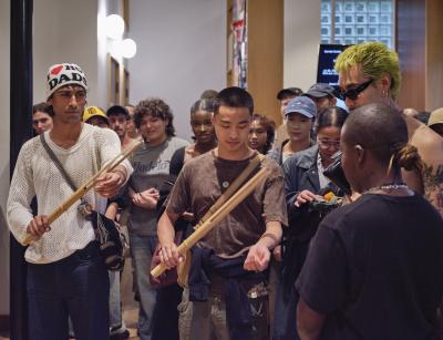 Two people stand each holding a bamboo buzzer in their left hand. The subjects both have their right arm out in front of them, as if they are banging the instrument on their arm to create noise. Tati au Miel stands in front of the two, watching them intently.