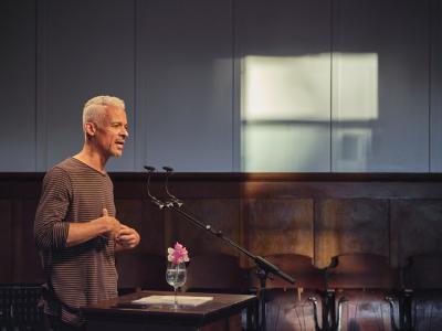Seen in profile, a poet with short white hair and salt-and-pepper stubble addresses a crowd from a lectern. He holds his hands by his sternum, giving the impression of both solemnity and earnestness. He wears a long-sleeve black and white striped shirt.