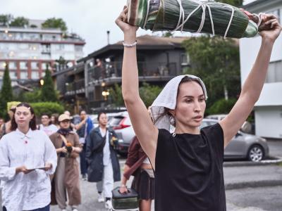 Bhenji Ra powerfully holds a wrapped and bound figurine of a saint above her head. She has a serious and stern look on her face. A group of people follow behind her.

