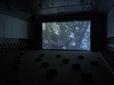 A projected image shows a person perched in a tree reaching with both hands for a green coconut wedged in the tree's limbs. They wear traditional Filipino garments.