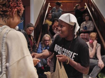 Tati au Miel wears a cloth atop their head like a bandana. They are wearing a black shirt that says IMPOSSIBLE in white, bold, uppercase letters. They are holding a brown bag in their right hand and are placing something in someone's hand with their left. The staircase behind them is crowded with people.