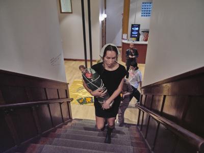 Bhenji Ra walks up the staircase at Western Front holding a figurine of a saint wrapped in palm leaves and bound with white rope. Performance attendees trickle in behind her.