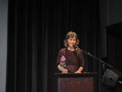 A poet with chin-length dark hair and bangs smiles as they address the crowd from a lectern, emanating warmth. She wears a brown loose knit sweater rolled up at the elbows, with a black tank top underneath.