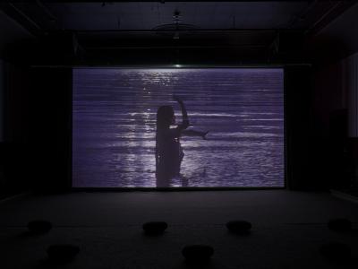 In a projected image we see a person waist deep in a body of water. Their silhouetted body forms an uppercase L with one arm reaching for the sky and the other one reaching along the horizon. Long, wet, hair lays stiff behind them. The sun is not present but is reflecting off of ripples on the water. 