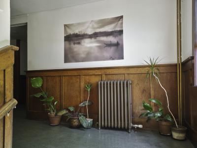 A sepia photograph printed on a sheer textile hangs at the top of the stairwell at Western Front. The image shows two boats distanced from one another on a body of water that is lined with trees. A vintage radiator and several potted plants are positioned on the floor below the print.