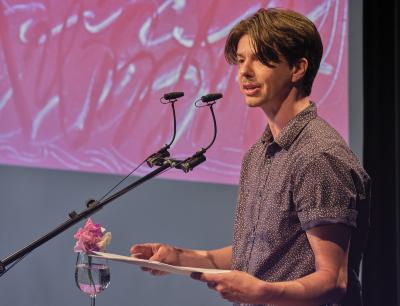 Seen in profile, a poet with short brown hair, a mustache, and a blue and white patterned shirt addresses a crowd from a lectern. They stand in front of a projection of red and white abstract art, holding a small stack of papers as they speak.