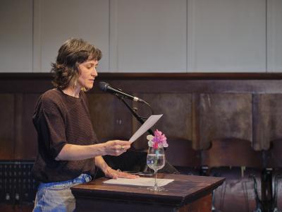 Side-angle view of a poet with chin-length dark hair wearing a brown knit sweater and jeans. She addresses an audience from behind a podium, looking down at a paper in their hand as she speaks. 